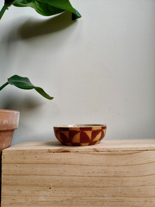 A small ceramic bowl with a brown and beige geometric pattern sits on a light wood block against a neutral wall, with a potted plant in the background.